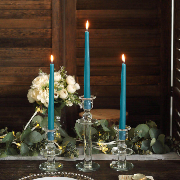 Three elegant teal taper candles in glass holders with flowers and greenery in the background.