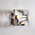 A contemporary polished stainless steel wall sconce with a clear glass lens and metallic frame, illuminated to showcase the lighting effect.