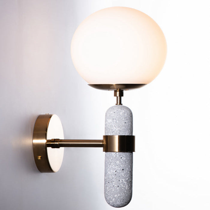 A modern wall sconce featuring a faux concrete body with a satin brass finish on the backplate and opal glass globe.