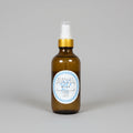 An amber glass room spray bottle with a white spray top and a gold collar, featuring a circular white label with blue text that reads 'BLUELILY ROOM SPRAY' and 'BLUE'.