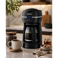 Catalog scene of black Cuisinart DCC-1500FR coffee maker with filled glass carafe, coffee cup, beans, honey, and sugar on a kitchen counter.